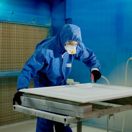 Man in protective workwear spraying paint on a piece of furniture at woodworking production facility.